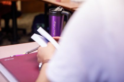 Student holding a book at a desk in a classroom setting.