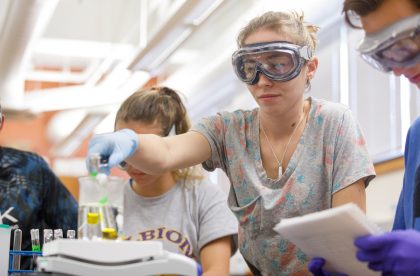 Students wearing protective goggles in a lab setting holding test tubes.