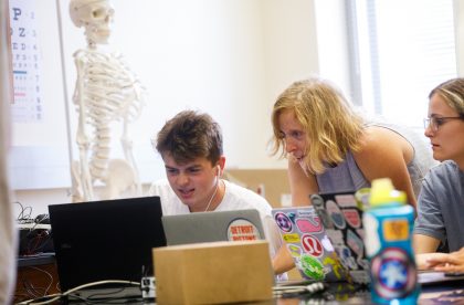 Students on computers in a lab setting, with a model of a skeleton in the background.