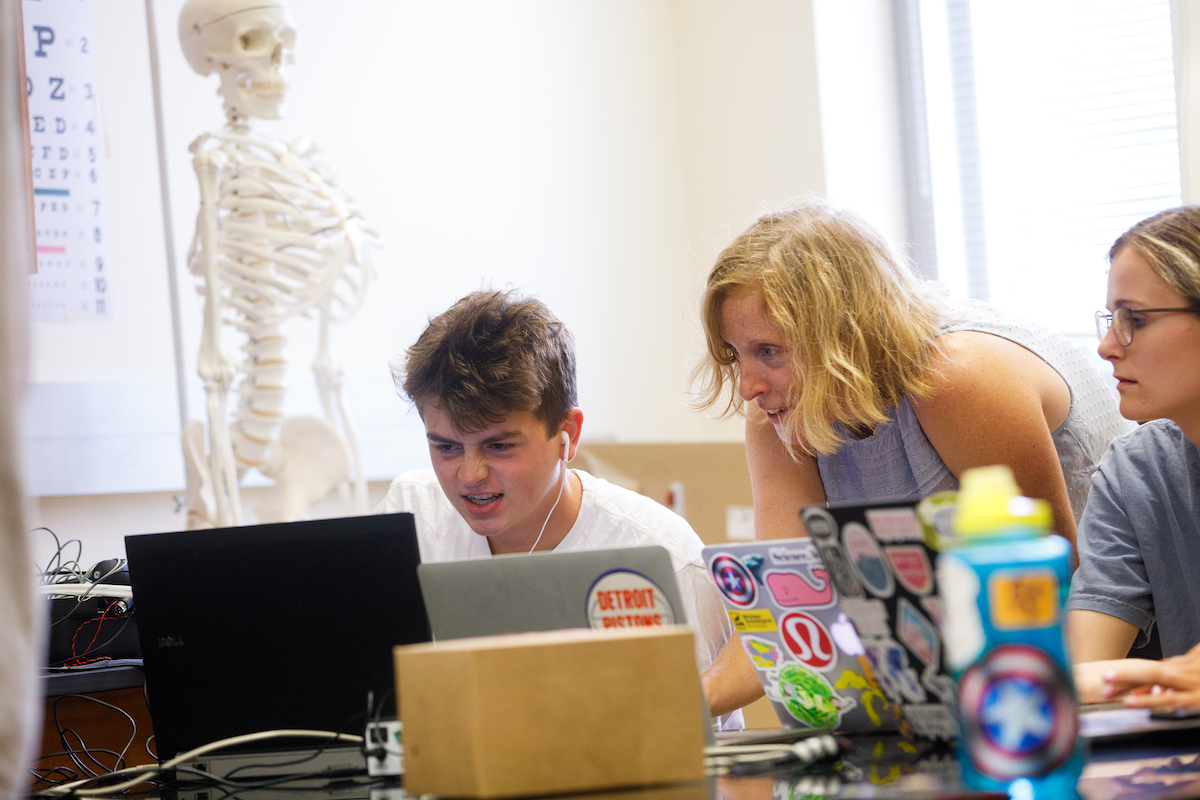 Students on computers in a lab setting, with a model of a skeleton in the background.