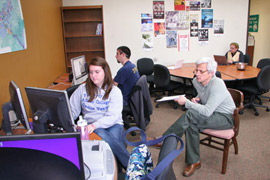 A woman sits at a desk in front of a computer. An older man sits in a chair next to her with his legs crossed. Two people sit behind them and desks in front of laptops. Posters decorate the back wall.