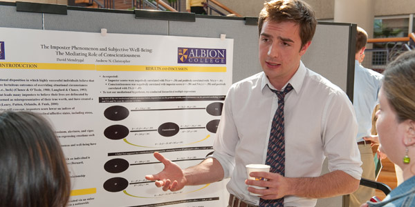 A man standing in front of a poster, talking and gesturing with his hands.