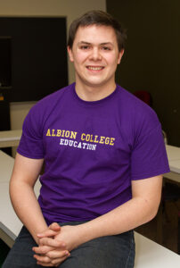 A male student wearing a purple shirt, smiling, with his hands folded in his lap.