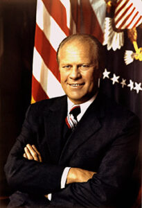 Photo of Gerald Ford in front of a flag. He is smiling and his arms are crossed in front of his chest.