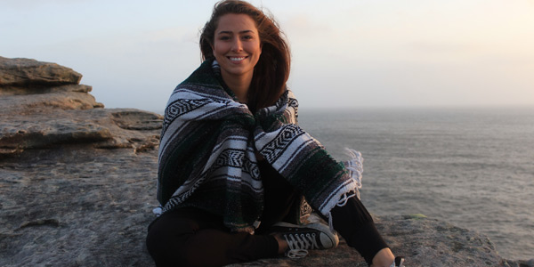 A student sits on the beach wrapped in a blanket. She is smiling at the camera.