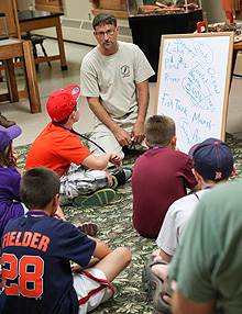 A man sitting on the floor surrounded by a group of children. Propped next to him is a white board.