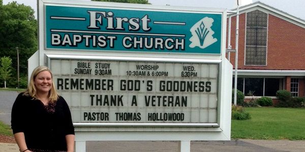 A student standing in front of a sign that reads: "First Baptist Church, Remember God's Goodness, Thank a Veteran"