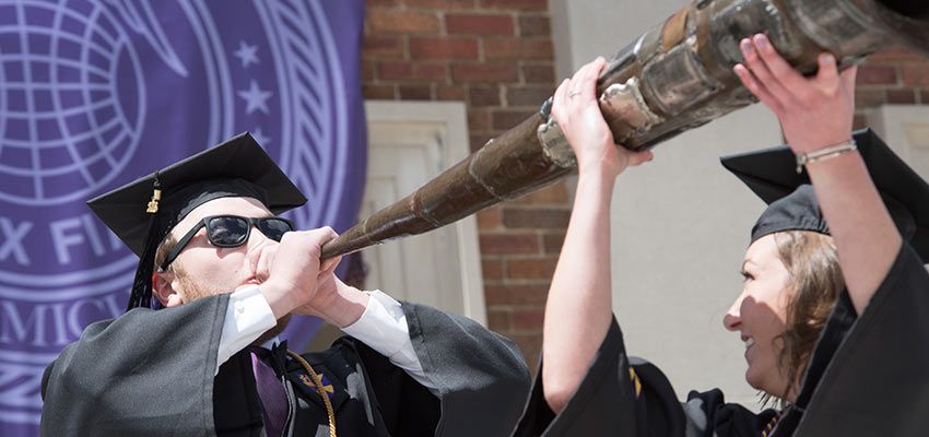 A student blowing into a large instrument while another student holds it up.