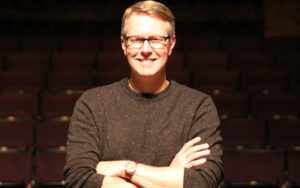 A professor smiling at the camera with his arms crossed. He is standing in an auditorium.