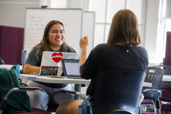 Two students facing each other with laptops in front of them.