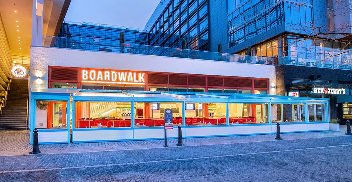 The front of Boardwalk is white with red trimmed windows, a lit sign reading Boardwalk, outdoor covered seating and a rooftop seating area. 