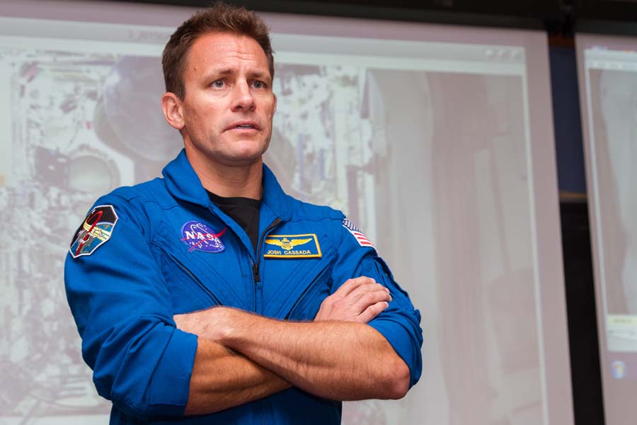 Josh Cassada wearing NASA flight suit coveralls stands with his arms crossed. Behind him a presentation on the International Space Station can be seen.