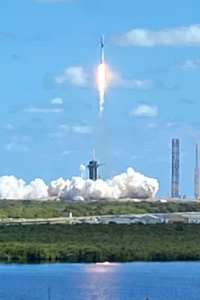 The Crew 5 rocket is lifting off the launch pad into a blue sky, as seen across a body of water. 