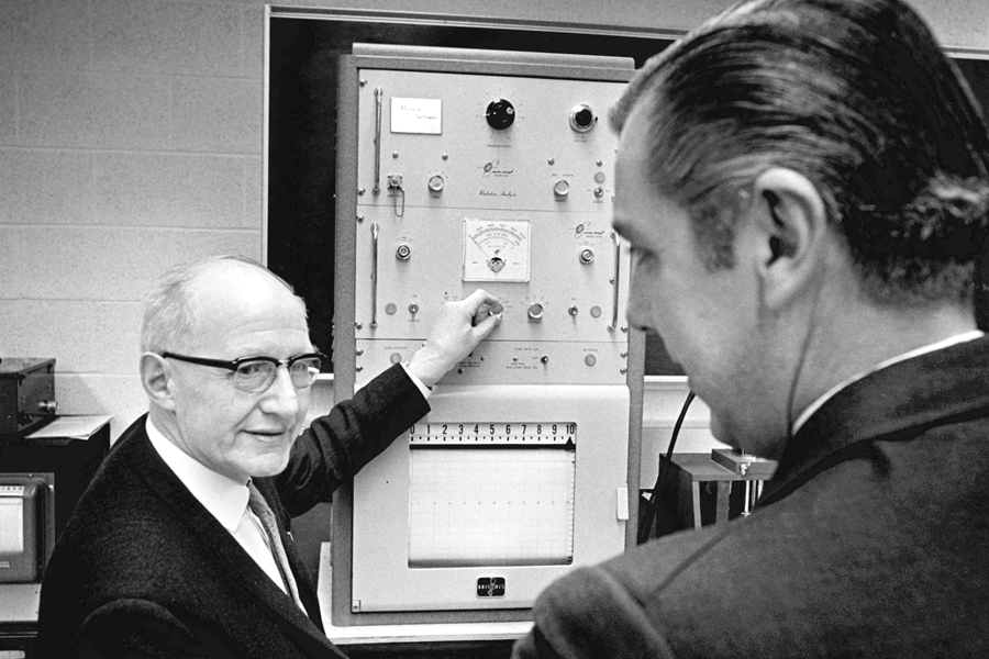 E.T.S. Walton is turning the knob of a scientific looking machine while he talks to a man in the foreground.