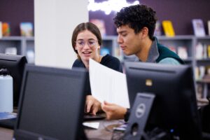 two students sitting in a computer lab talking together while one holds a sheet of paper
