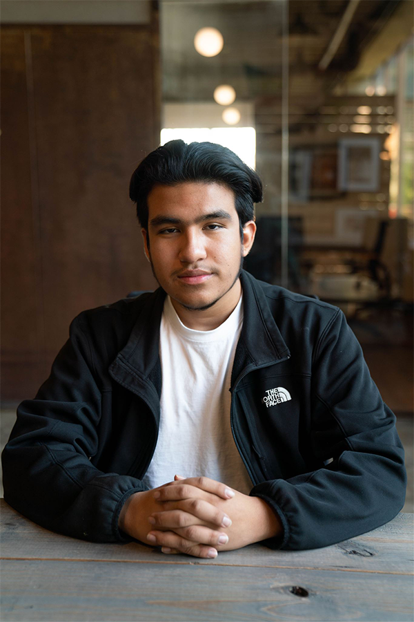 Adrian Sanchez sits at a wood table looking at the camera and has his hands clasped together. He is wearing a black North Face jacket and a white t-shirt. 