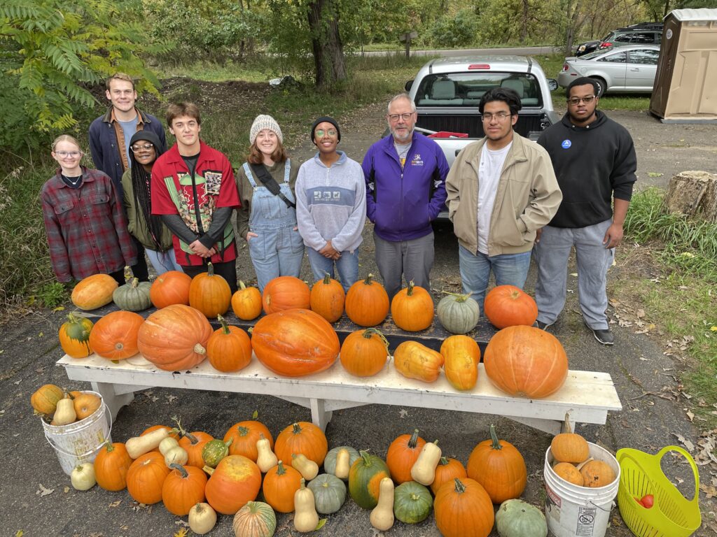 A group of CSE students standing behind a bench stacked with bright orange pumpkins and yellow squash